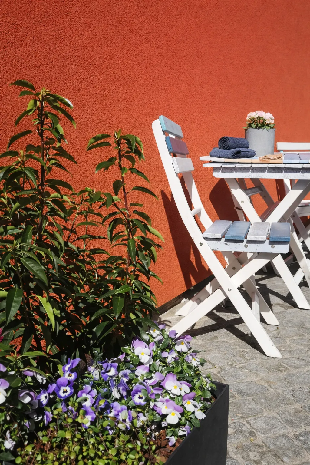 Detailaufnahme der Terrasse – weiße Klappstühle, Stiefmütterchen und Grünpflanzen vor orangefarbener Hauswand